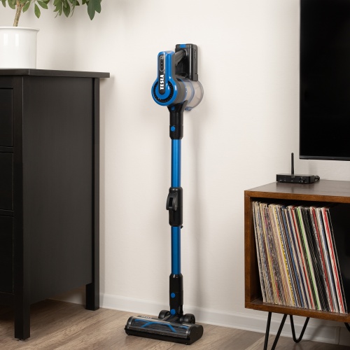 Vysavač TESLA Purestar E70 Aqua Pro parkovaný ve stojanu vedle knihovny v minimalistickém interiéru.