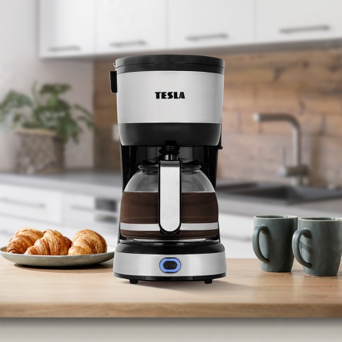Přední pohled na kávovar TESLA Coffeemaster ES200 s otevřeným víkem, vedle pečivo a dva šálky na kuchyňské lince.