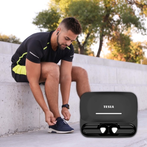 Muž sedící na schodech, sportovně oblečený, zavazuje si tkaničky a v uších má bezdrátová Bluetooth sluchátka TESLA Sound EB20 Black Raven. V popředí vpravo je nabíjecí pouzdro s logem TESLA.